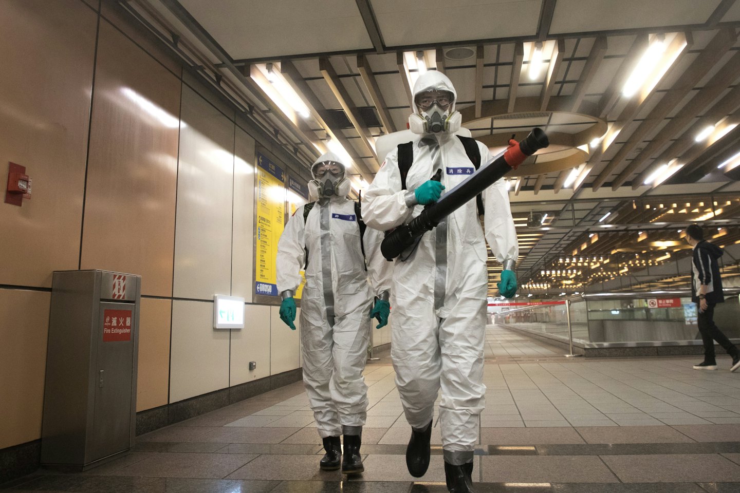 Two sanitation workers in hazmat suits walk through a subway station.